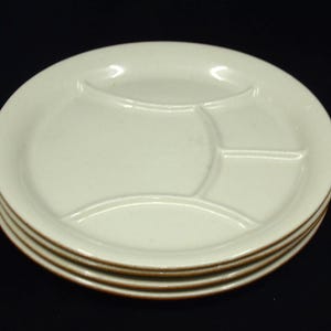 May include: Four off-white divided plates are stacked. Each plate has three sections and a brown rim. The plates are stacked against a black background. The plates are made of ceramic. The plates are approximately 25 cm in diameter.