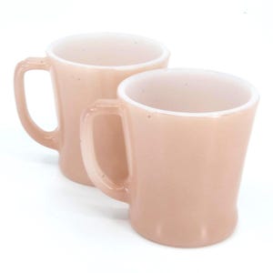Pode incluir: Duas canecas vintage de vidro rosa com interiores brancos. Cada caneca tem uma alça curva e uma forma ligeiramente alargada. As canecas estão sobre um fundo branco, destacando sua cor pastel suave e design retrô.