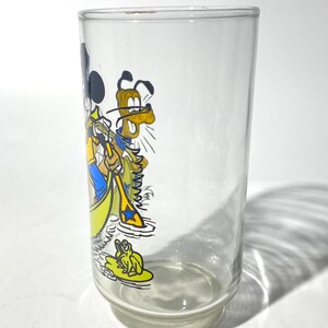 May include: A clear glass tumbler featuring a colorful cartoon illustration of Mickey Mouse and Pluto in a yellow boat. The glass has a wide base and a straight, cylindrical body. The cartoon characters are depicted in bright colors.