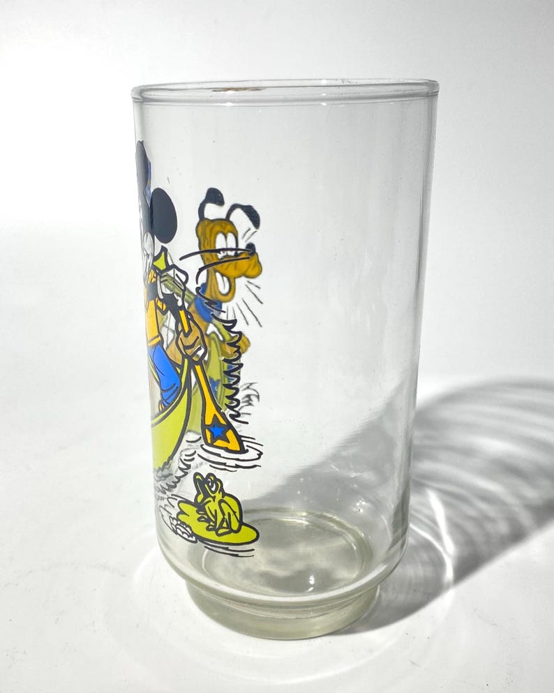 May include: A clear glass tumbler featuring a colorful cartoon illustration of Mickey Mouse and Pluto in a yellow boat. The glass has a wide base and a straight, cylindrical body. The cartoon characters are depicted in bright colors.