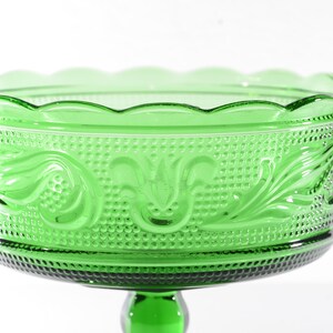 May include: A green glass pedestal bowl with a scalloped rim and a decorative floral pattern. The bowl is on a pedestal with a round base.