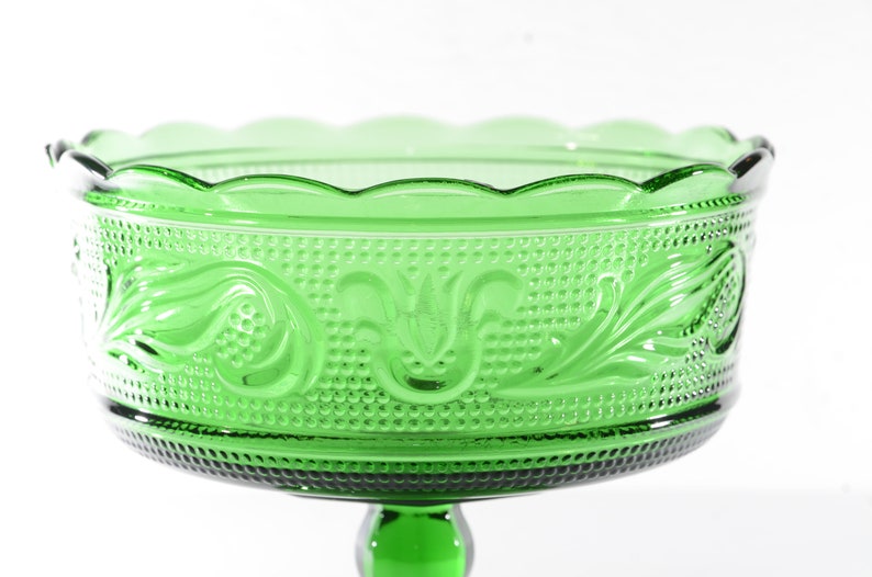 May include: A green glass pedestal bowl with a scalloped rim and a decorative floral pattern. The bowl is on a pedestal with a round base.