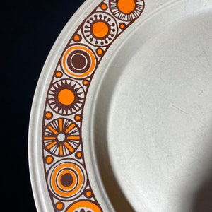 May include: A white ceramic plate with a brown and orange geometric design around the edge. The design features circles, stars, and flowers.