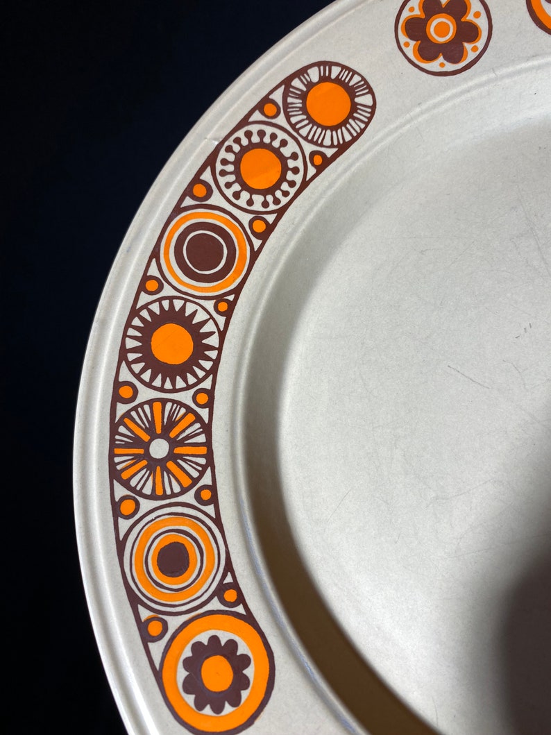 May include: A white ceramic plate with a brown and orange geometric design around the edge. The design features circles, stars, and flowers.