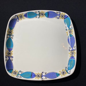 Può includere: Un piatto quadrato in ceramica bianca con un design di pesci blu e dorati attorno al bordo.