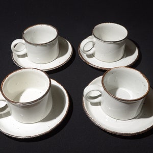 May include: Set of four white ceramic coffee cups with brown rims and speckles. Each cup is paired with a matching saucer.