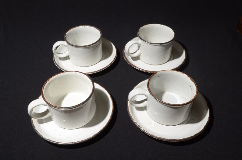 May include: Set of four white ceramic coffee cups with brown rims and speckles. Each cup is paired with a matching saucer.