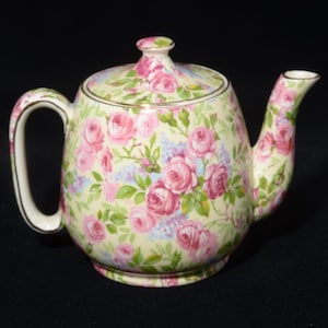 May include: A porcelain teapot featuring a cream-coloured base adorned with a dense pattern of pink roses, green leaves, and small blue floral accents. The teapot includes a matching lid, a curved handle, a spout, and gold-toned trim on the edges.