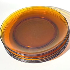 May include: A stack of four round, translucent amber-coloured glass plates. Each plate features a smooth, circular surface with a slightly raised rim, showcasing a warm, golden-brown hue that allows light to pass through the material.