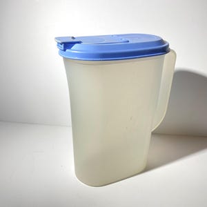 Puede incluir: Una jarra de plástico transparente con tapa azul y asa. La jarra está vacía.