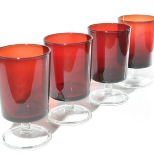 May include: Four red-tinted glass goblets with clear stems and bases. The glasses have a cylindrical shape and are arranged in a row against a white background. The red colour is most intense at the top of the glasses.