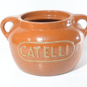 Op de afbeelding: Een terracottakleurige keramische pot met twee handvatten en een deksel. De pot heeft een wit ovaal etiket met het woord "CATELLI" in zwart gedrukt.