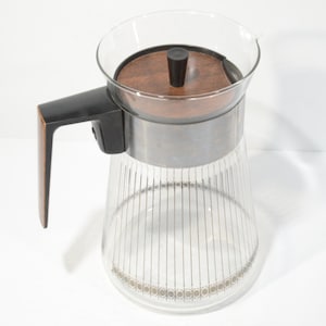 Op de afbeelding: Een glazen koffiekan met een houten deksel en een zwarte handgreep. De kan heeft een zilveren band rond het midden en een goud-wit patroon rond de bodem.