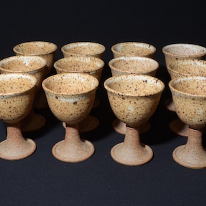 Puede incluir: Un juego de doce copas de cerámica moteada. Las copas tienen una base marrón claro y un cuenco moteado de color beige. El tallo y la base son de color marrón rojizo. Las copas están dispuestas en tres filas de cuatro.