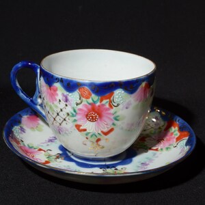 Op de afbeelding: Een wit theekopje en schotel met een blauwe rand en een bloemenpatroon. Het kopje heeft een handvat en de schotel heeft een gegolfde rand.