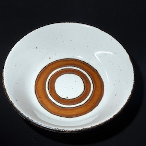 May include: A white ceramic bowl with a brown concentric circle design in the center. The bowl has a speckled texture and a dark brown rim. The bowl is set against a black background.