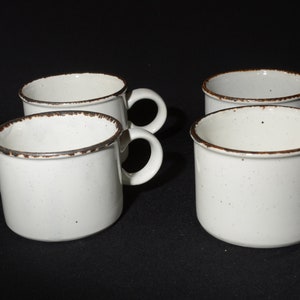 May include: Four white ceramic mugs with brown rims. The mugs have a speckled glaze and are arranged in a row on a black surface.