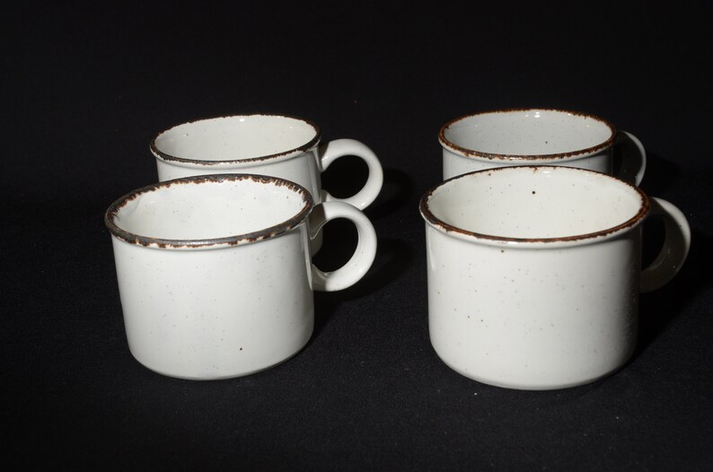 May include: Four white ceramic mugs with brown rims. The mugs have a speckled glaze and are arranged in a row on a black surface.