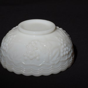 K&ouml;nnte beinhalten: Eine wei&szlig;e Schale im Vintage-Stil mit einem detaillierten Trauben- und Bl&auml;ttermuster. Die Schale hat einen gewellten Rand und einen kleinen, kreisf&ouml;rmigen Sockel. Die Schale besteht aus undurchsichtigem wei&szlig;em Glas und steht vor einem schwarzen Hintergrund.