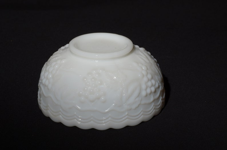 K&ouml;nnte beinhalten: Eine wei&szlig;e Schale im Vintage-Stil mit einem detaillierten Trauben- und Bl&auml;ttermuster. Die Schale hat einen gewellten Rand und einen kleinen, kreisf&ouml;rmigen Sockel. Die Schale besteht aus undurchsichtigem wei&szlig;em Glas und steht vor einem schwarzen Hintergrund.