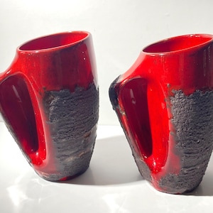 Pode incluir: Duas canecas de cerâmica vermelha com um design único. A metade superior de cada caneca é vermelha brilhante, enquanto a metade inferior apresenta uma aparência texturizada, marrom escuro, quase vulcânica. Cada caneca tem uma alça grande e aberta.