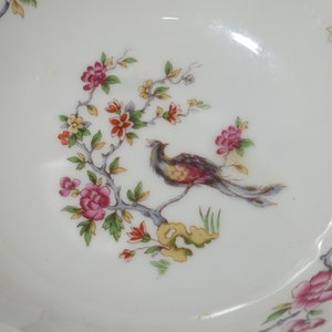 May include: A white porcelain bowl with a gold rim features a delicate floral design with a bird perched on a branch. The flowers are pink, red, and yellow, and the bird is brown, red, and yellow.