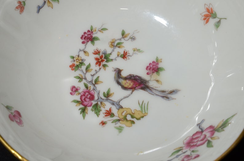 May include: A white porcelain bowl with a gold rim features a delicate floral design with a bird perched on a branch. The flowers are pink, red, and yellow, and the bird is brown, red, and yellow.