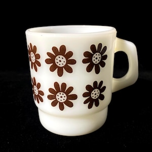 Op de afbeelding: Een witte melkglazen mok met een herhalend patroon van bruine madeliefachtige bloemen met gestippelde harten. De mok heeft een cilindervormig lichaam met een getrapte voet en een stevig, geïntegreerd handvat aan de rechterkant voor warme dranken.