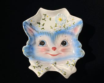 Könnte beinhalten: Eine weiße Keramikschale in Form eines Katzengesichts mit blauen Fellakzenten, großen dunklen Augen, einer rosa Nase und einem lächelnden Mund. Die Schale ist mit gemalten blauen und weißen Blumen auf der Stirn und einer Schleife am Kinn verziert.