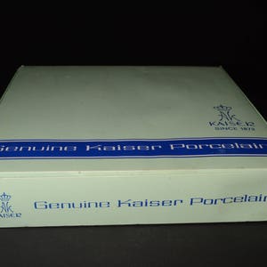 May include: A rectangular box with a pale green lid and blue text reading "Genuine Kaiser Porcelain." The box features the Kaiser logo and the text "SINCE 1872." The box is set against a black backdrop.