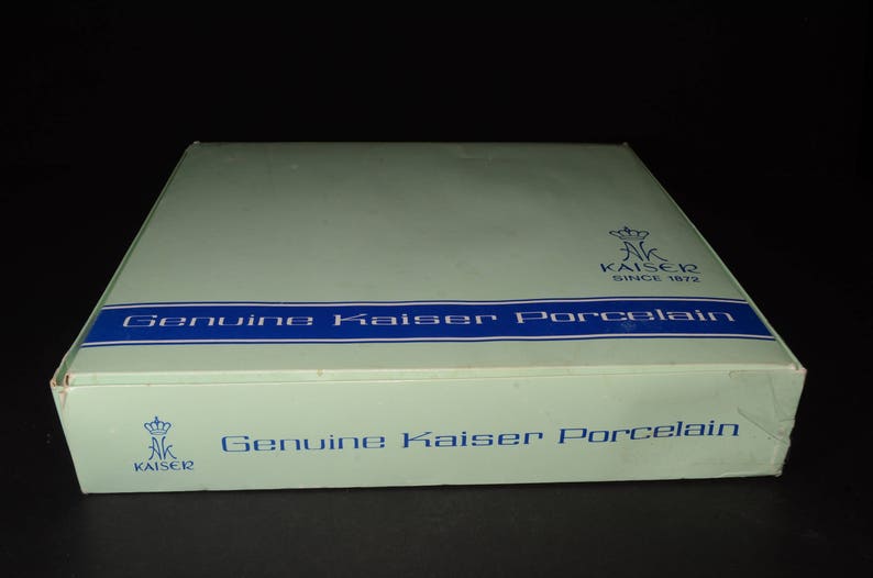 May include: A rectangular box with a pale green lid and blue text reading "Genuine Kaiser Porcelain." The box features the Kaiser logo and the text "SINCE 1872." The box is set against a black backdrop.