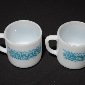 May include: A pair of white ceramic mugs, each featuring a turquoise floral pattern around the base and a curved handle. The mugs are displayed on a black surface, highlighting their simple design.