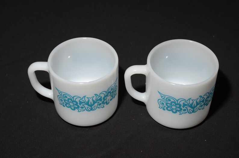 May include: A pair of white ceramic mugs, each featuring a turquoise floral pattern around the base and a curved handle. The mugs are displayed on a black surface, highlighting their simple design.
