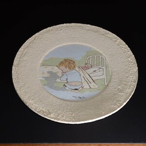 May include: A white ceramic plate with a raised rim features a hand-painted illustration of a child in blue pajamas praying beside a bed with a teddy bear inside. The illustration is set against a light blue background with a green grassy area.