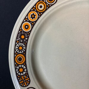 May include: A white ceramic plate with a brown and orange geometric pattern around the edge.