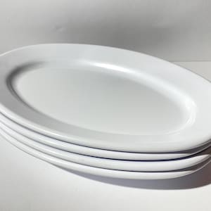 Puede incluir: Una pila de cuatro fuentes ovaladas blancas. Los platos están hechos de un material brillante y tienen un diseño simple y elegante. Los platos están apilados uno encima del otro, con el plato superior ligeramente inclinado.