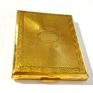 Könnte beinhalten: Ein rechteckiges Kompaktetui aus goldfarbenem Metall mit einem Strahlenmuster, das von einer leeren rechteckigen Platte in der Mitte ausgeht. Der Rand ist mit einem geprägten Blattmotiv verziert und das Etui hat einen kleinen Verschluss.