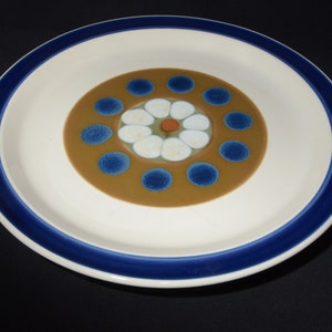 Puede incluir: Un plato de cerámica blanco con un borde azul y un centro marrón con un diseño de flor blanca y puntos azules.