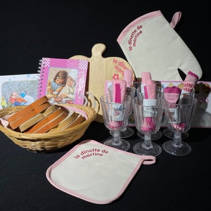 May include: A pink and white play kitchen set with a cookbook, a wooden cutting board, a pot holder, a basket of bread, and six glasses with pink napkins. The set includes the text "la dinette de martine" on the pot holder and the cookbook.