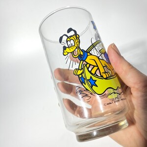 May include: Clear glass tumbler featuring a cartoon image of Pluto in a yellow and blue design. The glass has a cylindrical shape with a slightly flared base. The design includes the text "WALT DISNEY PRODUCTIONS".