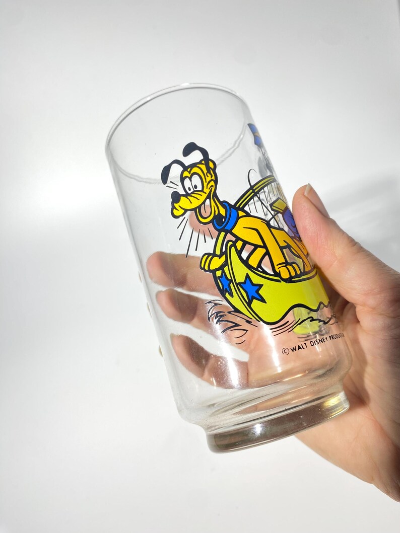 May include: Clear glass tumbler featuring a cartoon image of Pluto in a yellow and blue design. The glass has a cylindrical shape with a slightly flared base. The design includes the text "WALT DISNEY PRODUCTIONS".