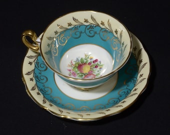 May include: A teal and gold teacup and saucer set with a floral and fruit design. The saucer has a scalloped edge.