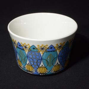 Può includere: Una ciotola in ceramica bianca con un design di pesci blu e gialli. La ciotola ha una forma arrotondata e un bordo largo.