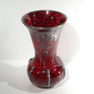 Puede incluir: Un jarrón de vidrio rojo con detalles plateados. El jarrón tiene una base redonda y un cuello estrecho. Los detalles plateados tienen forma de diseño floral.