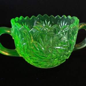 May include: A green glass sugar bowl with two handles. The bowl has a cut glass design and is made of uranium glass, which glows under a black light.