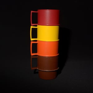 May include: Four brightly coloured plastic mugs stacked on top of each other. The mugs are in shades of red, yellow, orange, and brown. Each mug has a rectangular handle. The mugs are set against a black background.