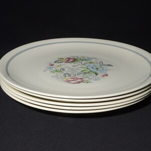 May include: A stack of five white ceramic dinner plates with a blue rim and a floral design in the centre. The flowers are pink, blue, yellow, and green.