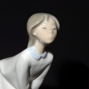 May include: A porcelain figurine of a young girl with long blonde hair wearing a white dress with a blue collar. The girl is looking down with a serious expression.