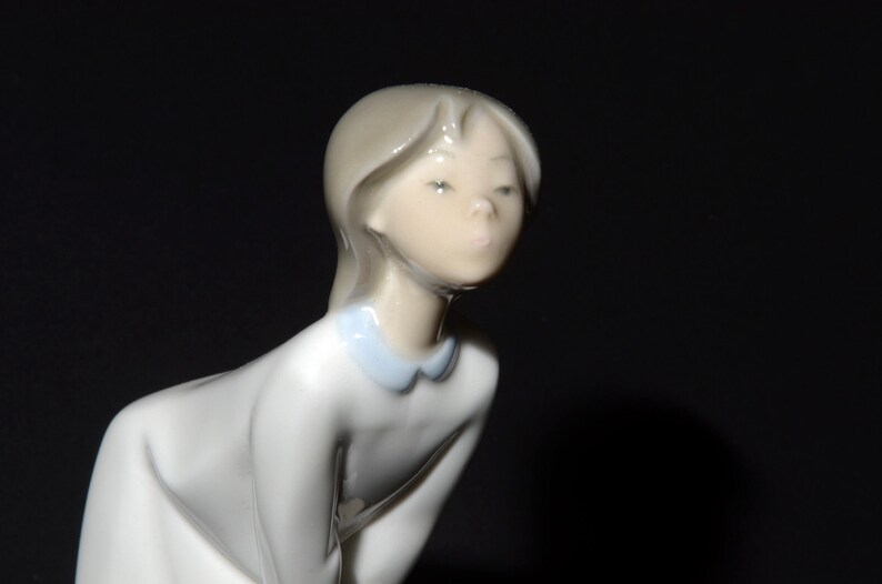 May include: A porcelain figurine of a young girl with long blonde hair wearing a white dress with a blue collar. The girl is looking down with a serious expression.