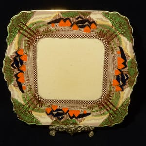 Może przedstawiać: Kwadratowy ceramiczny talerz do serwowania z kremowym środkiem i dekoracyjną obwódką. Obwódka przedstawia krajobraz z pomarańczowymi budynkami, zielonymi drzewami i wzorem w szachownicę. Talerz jest podparty ozdobnym metalowym stojakiem.